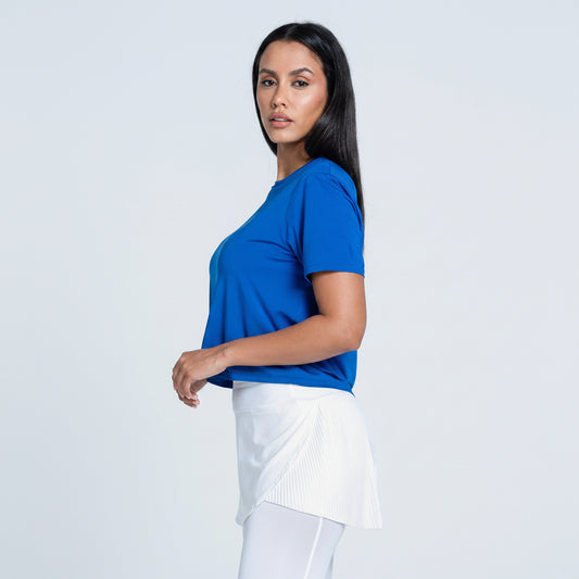 Side view of a woman wearing a blue boxy crew neck dri-fit cropped performance t paired with a white tennis skirt.