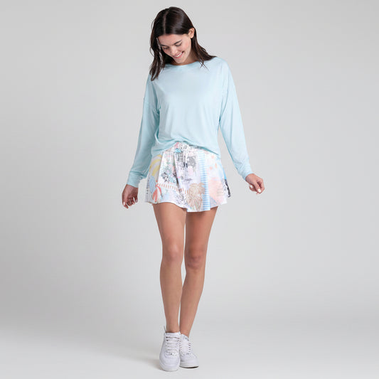 Young woman wearing a light blue long-sleeve top and pastel patterned tennis skirt on a white background