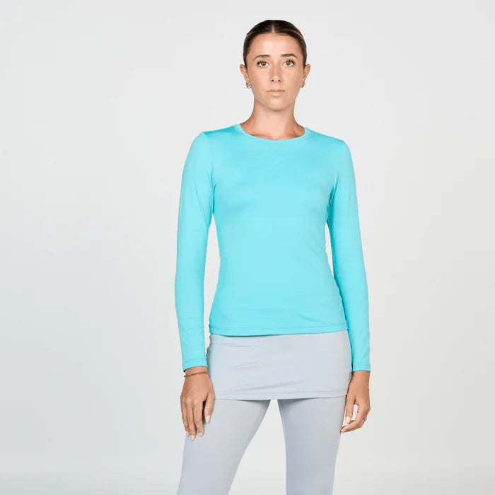 A woman wearing a long sleeve blue tennis top and light gray tennis skirt with built in capri pants, standing against a white background.