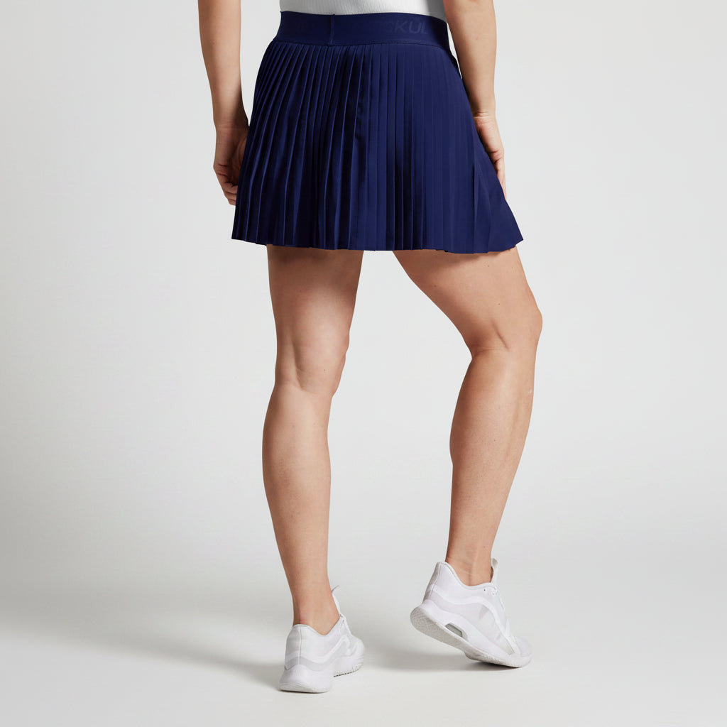 Woman wearing a navy blue pleated tennis skirt and white sneakers on a gray background