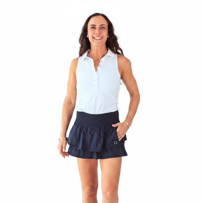 Woman wearing a white sleeveless shirt and navy tennis skirt against a light blue background