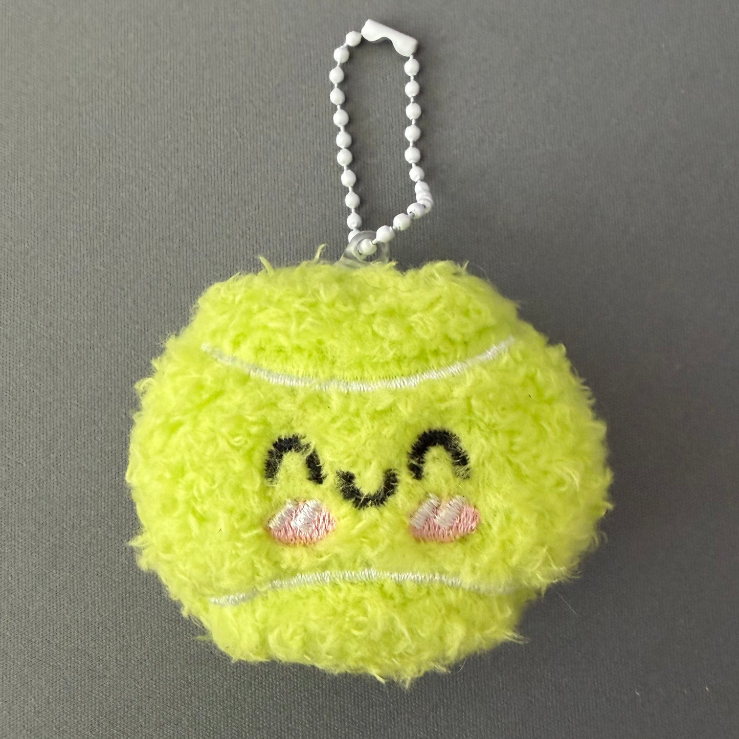 Yellow fuzzy keychain shaped like a tennis ball with decorative elements on a gray background