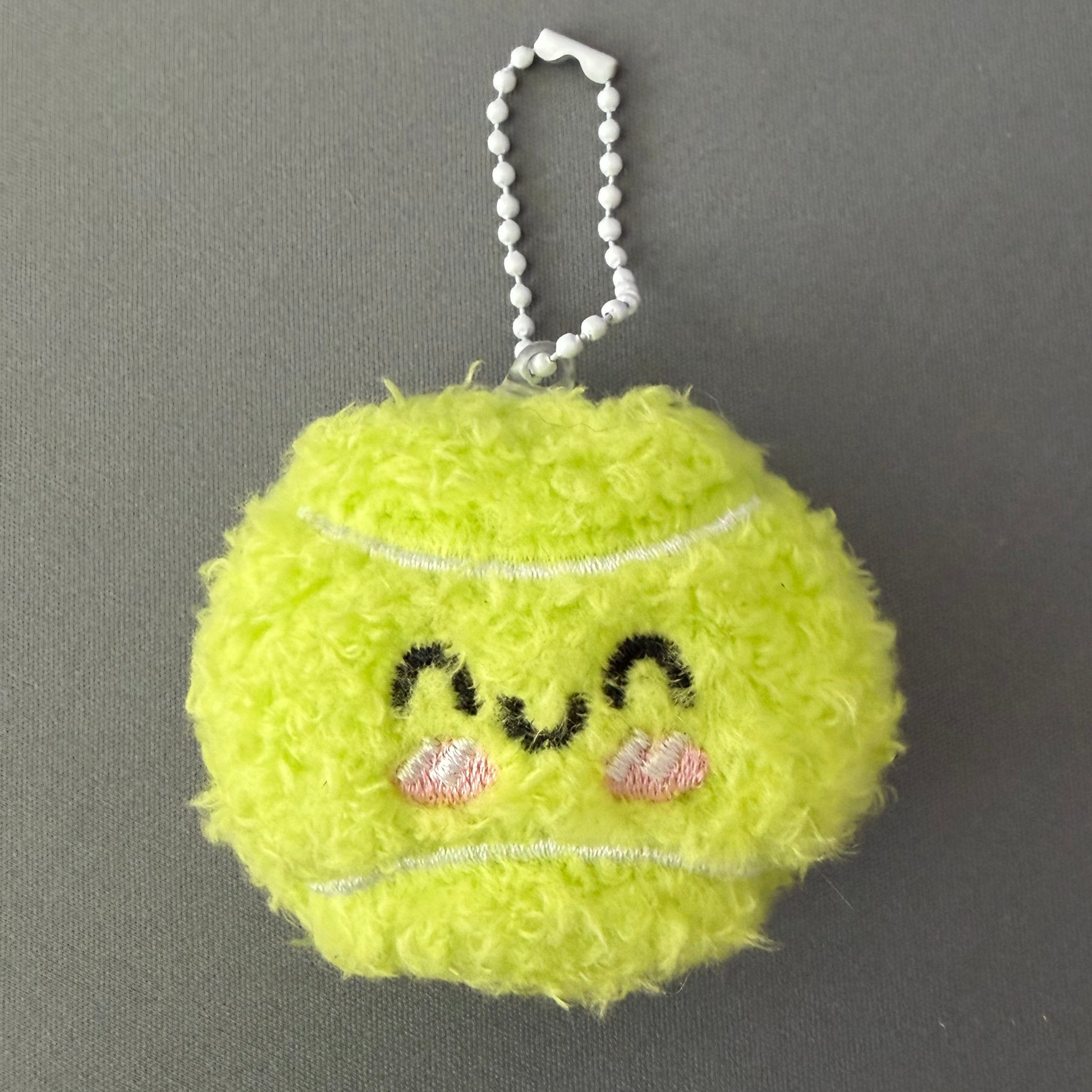 Yellow fuzzy keychain shaped like a tennis ball with decorative elements on a gray background