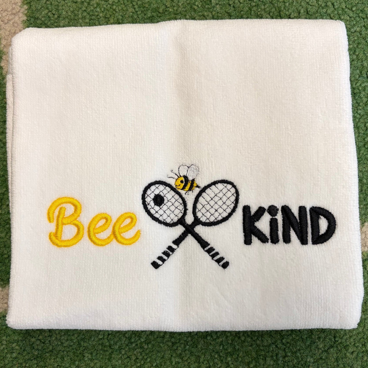 White towel embroidered with yellow Bee, a small bee, crossed racquets, and black KIND lettering