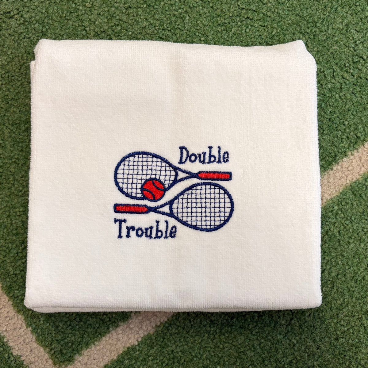 White towel embroidered with Double Trouble, tennis rackets, and a red ball